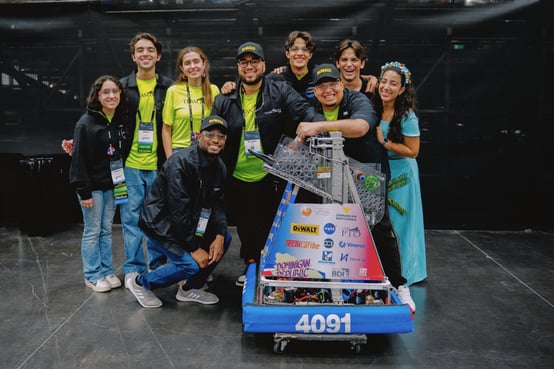 Enmanuel stands in a group with his student FRC team. They are all smiling big and surrounding their robot.