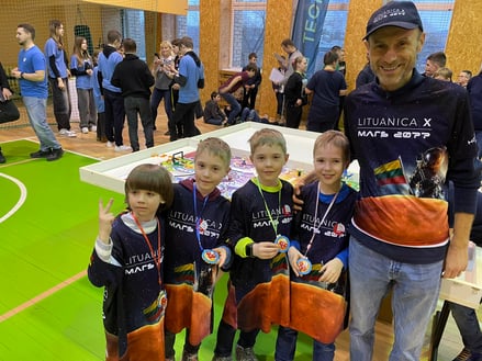 Algirdas poses with four young members of his FIRST LEGO League team. They are smiling and excited at a competition and are wearing team gear with an image of the Lithuania flag on Mars.