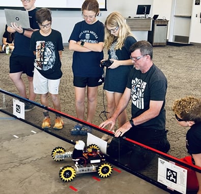Brian kneels next to a FTC field, instructing his students. Their robot sits in front of them, and they are getting ready to drive.