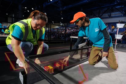 Two FTC volunteers bend over the FTC field, inspecting it and making sure everything is set up and in order. They are wearing FIRST volunteer shirts and safety protection.