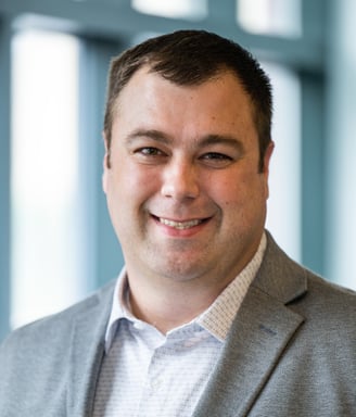 Michael smiles and poses for a professional headshot. He is wearing a gray suit and a patterned white button up.