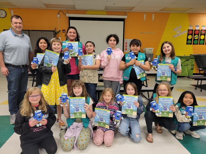 Michael poses with a group of Girl Scouts holding the "Life of a Can" book. They are all smiling and holding a small plushie of the can character from the book. They are in a classroom.