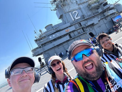 Jon takes a selfie with other FIRST referees. They are smiling and posing in front of a large Navy ship. He is wearing the inclusive Pride flag FIRST referee jersey.