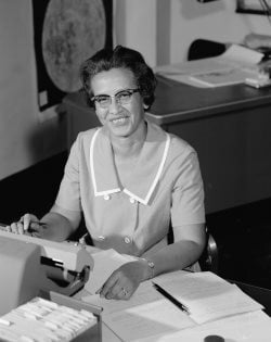 Picture of Dr. Katherine Johnson hard at work at her desk, smiling. The image is in black and white.