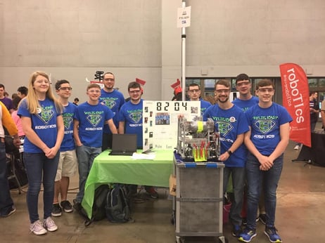 Lauren as a student with her FTC team. They are standing near their presentation, wearing safety glasses, and smiling.