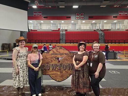 Lauren poses with other volunteers in front of a western themed FIRST sign. They are dressed as cowgirls.
