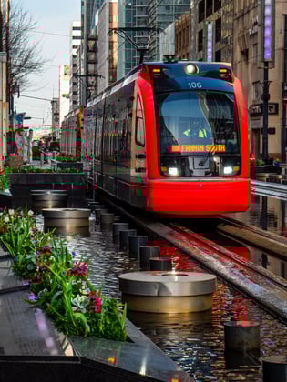 A red metro rail car drives down Main Street in Houston, Texas. Flowers line the streets and water features can be seen around the rail line.