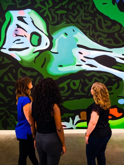 A group of people look at a brightly lit and colorful art piece. The art piece doesn't resemble anything specific, and is stylistic with chunky lines. The main outer color focus is on green, and the inner color focus is light blue.
