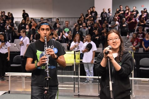 Sarah and her teammate stand performing the National Anthem with a crowd of people behind them. Sarah is holding the microphone singing and her teammate is playing the clarinet.