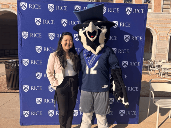 Sarah stands in front of a blue stand up background with the Rice University logo across the back. She is posed and smiling next to the Rice University mascot who is an owl dressed in a blue Rice sports uniform.