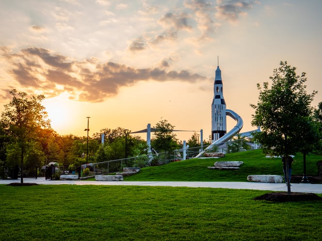 A space-themed playground can be seen at a grassy Hermann Park in Houston, Texas. The sun is setting in the distance.