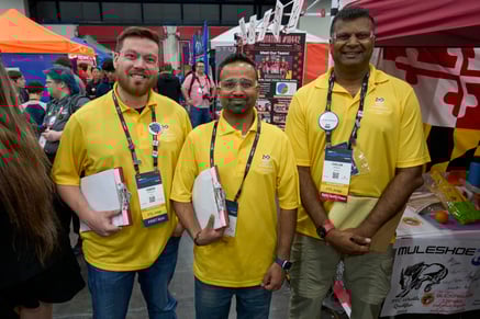 Three FIRST volunteers smile and pose for the camera in the FLL pits. They are holding their clipboards and walking around judging.