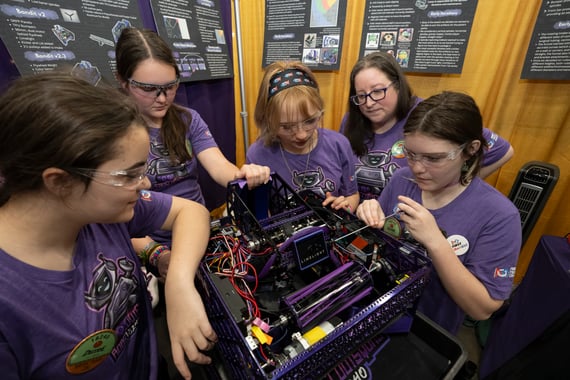 A group of female FTC students and their mentor gather around their robot, working on on it in their pit.