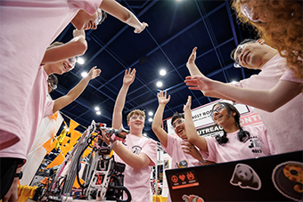 A group of FIRST students stand in a circle smiling, hands in the air, finishing a cheer.