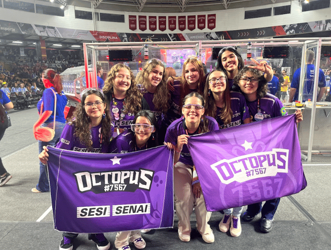 Luiza poses as a mentor of team "Octopus." They all smile, holding two purple banners with their team name and number.