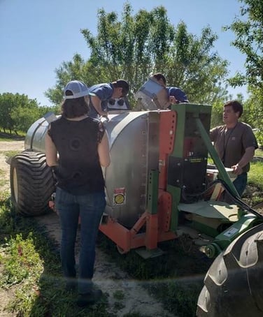 Megan stands in front of three people working on a tractor, observing their actions. She is facing away from the camera.