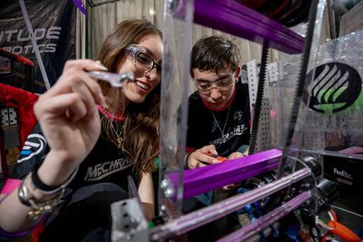 Two students smile and bend over their robot.. They are using a wrench to work on it and are wearing sunglasses.