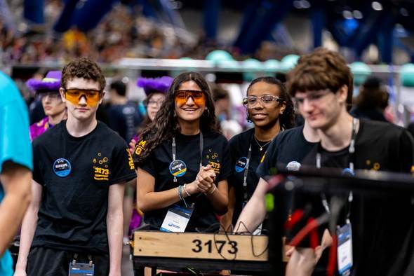 Four students stand watching an event. They are smiling and wearing safety glasses.