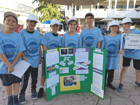 The Heschel RoboHawks pose with their impact project poster. They are smiling and wearing blue RoboHawks shirts.