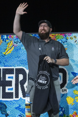 Cameron poses in front of the Submerged Logo display from 2025 FIRST Championship. He is holding his Volunteer of the Year award shirt and waving, smiling at the crowd.