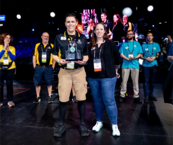 Chris poses with other FIRST referees and volunteers, holding his award and smiling for the camera as they clap.