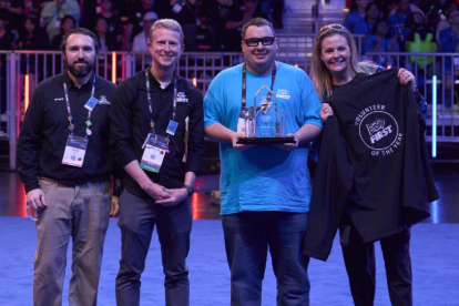 Thad poses for their award with other members of FIRST leadership. They are smiling wide and wearing safety glasses on the floor of FIRST Championship
