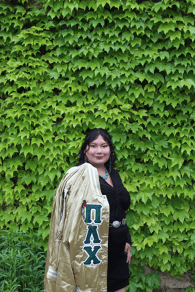 Alicia stands in her Sorority jacket in front of an ivy covered wall. She is smiling and dressed up.