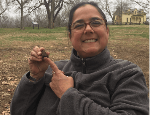 Mel smiles at a digsite, pointing to an artifact she found.