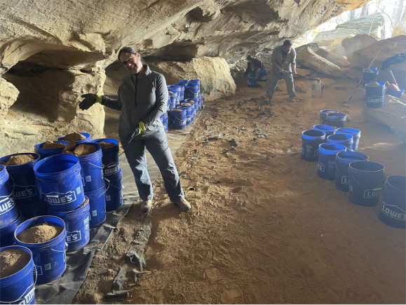 Mel smiles and gestures to Lowes buckets full of sand. She is standing in a cave where others can be seen in the background excavating.