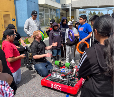 The lead mentor for the LHS Stingers gestures to their robot, explaining something as students look on.