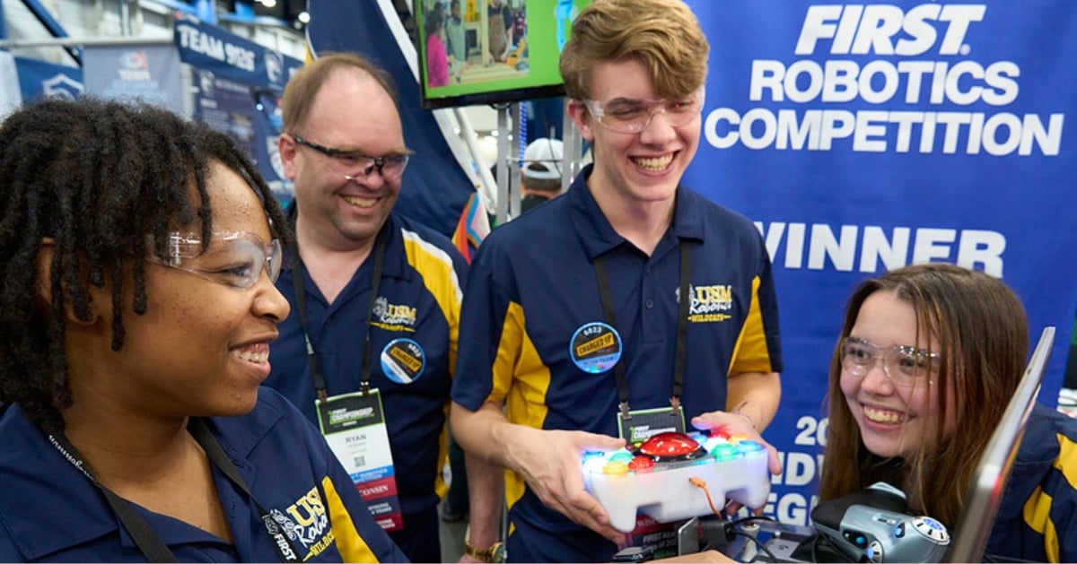 Students stand with their mentor around a computer and are smiling.