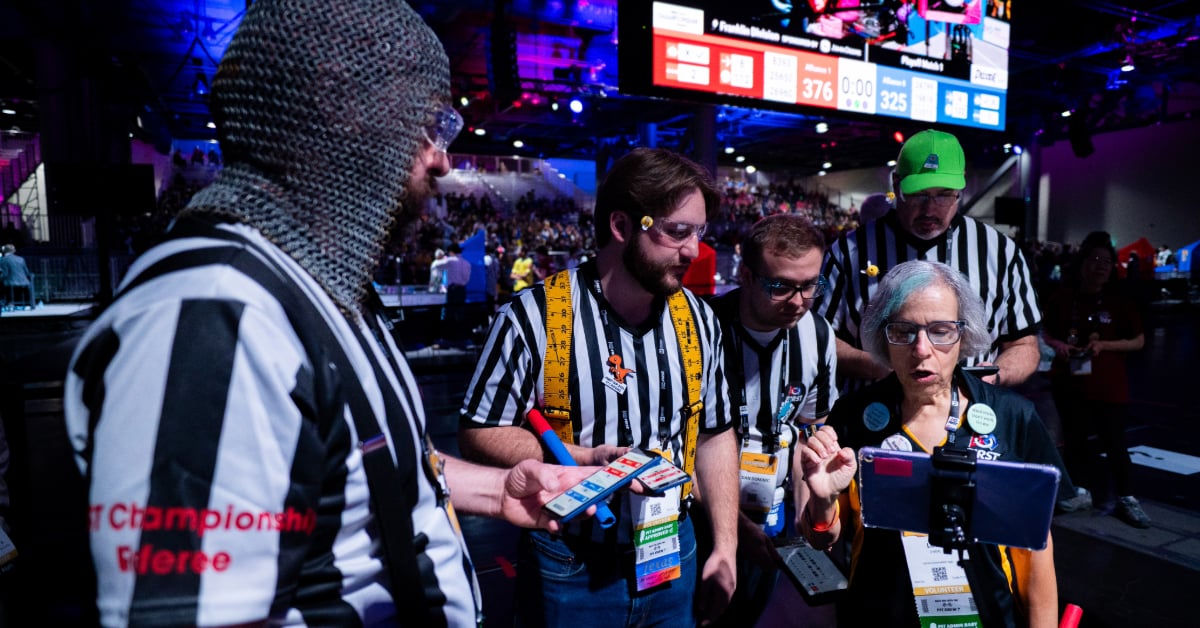 A group of referees all stand in a circle discussing a recent match. One is wearing chainmail on their head.
