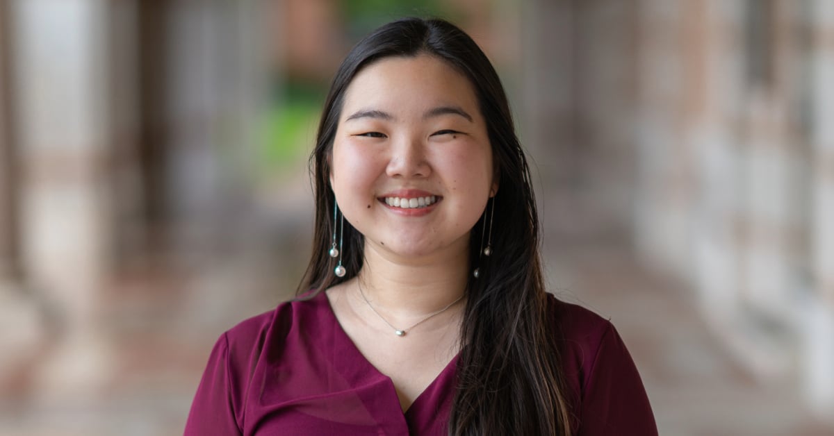 Sarah smiles for a professional headshot in front of a blurred outdoor background. She is wearing wearing a maroon blouse with dangly earrings and a small necklace.