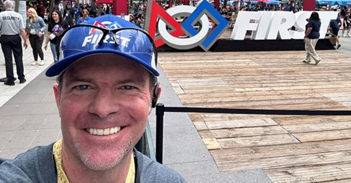 Brian takes a selfie with a big smile on his face in front of the FIRST sign at Championship. He is wearing a FIRST hat and safety glasses on top of his head.