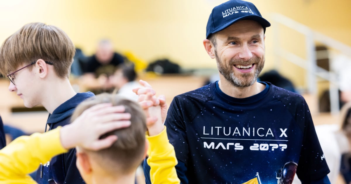 Algirdas smiles at a FIRST match. Two students can be seen to his left excitedly watching a match. 