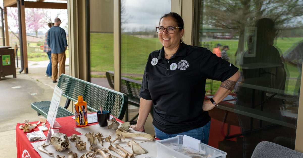 Mel smiles, standing in front of an archaeologic display table where she is presenting. The table is filled with bones and artifacts.