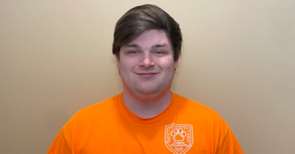 Ryan stands in front of a plain background, smiling for the camera. He is wearing an orange Team shirt.