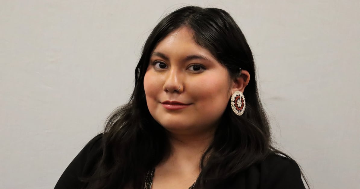 Alicia stands in front of a blank wall, smiling for a professional headshot. She is wearing traditional Indigenous-style earrings.