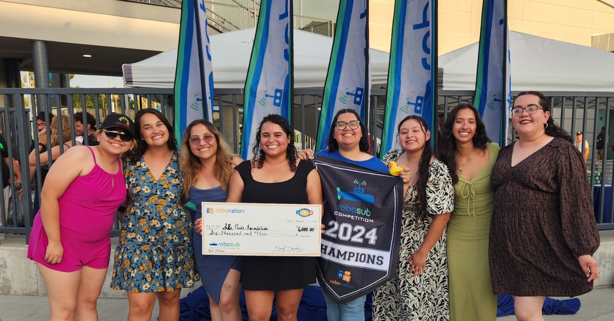 Team Desert WAVE smiles and poses in front of the RoboSub competition flags holding their first place prize in the form of a large check and a winning banner.