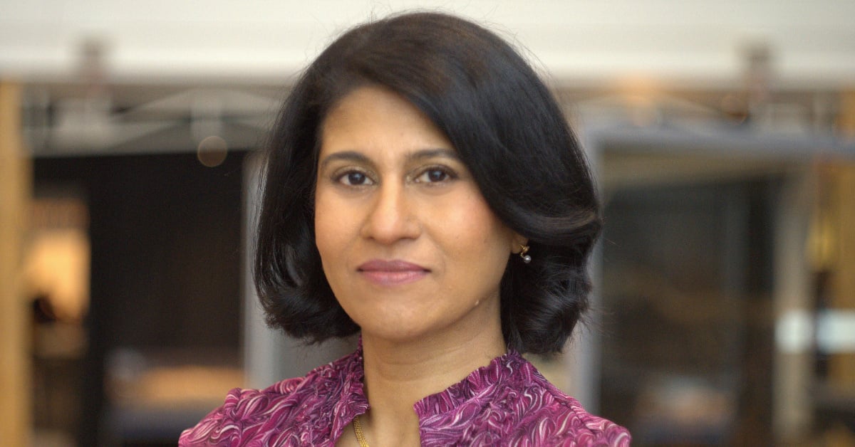 Jayshree Seth smiles for her professional headshot in front of a blurred office background. She is wearing a purple blouse.