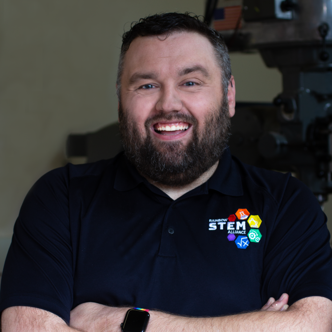 Jon is smiling wide with his arms folded. His shirt displays the Rainbow STEM Alliance logo.