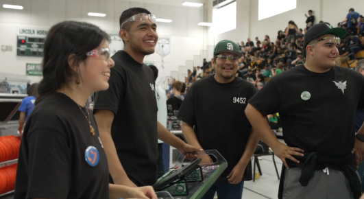 LHS Stingers students stand together smiling and laughing at a robotics competition. They are wearing safety goggles and team gear.