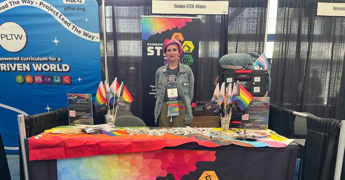 Rae stands behind the Rainbow Stem Alliance booth at Championship. They are surrounded in Pride flags and smiling big!
