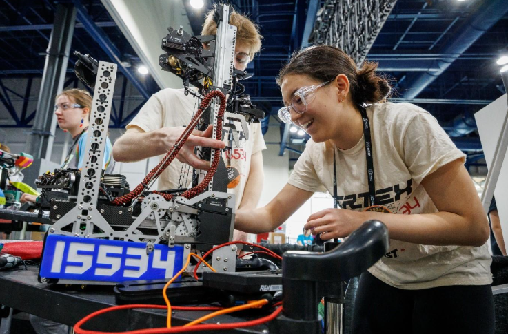 Two students wearing safety goggles smile as they lean over their robot, adjusting it.