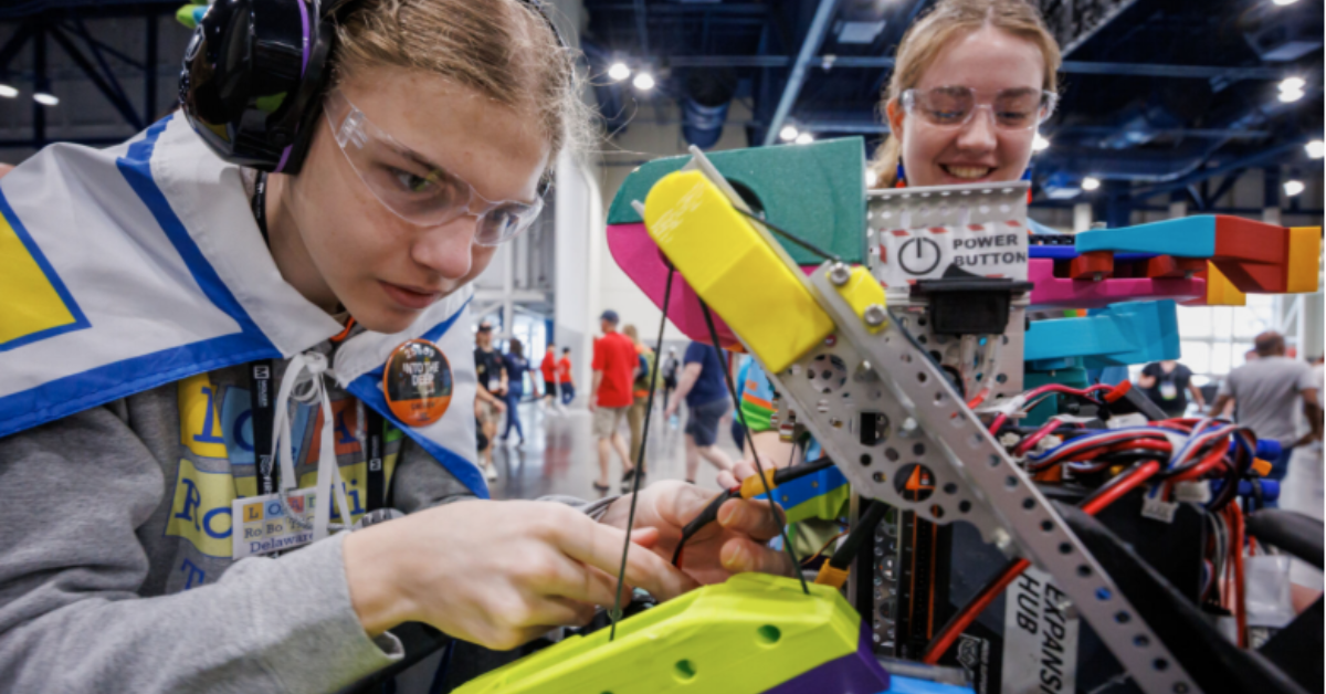 Two students focus on working on their robot. They are smiling and wearing safety goggles, with the student closest to the camera also wearing noise-cancelling ear muffs.